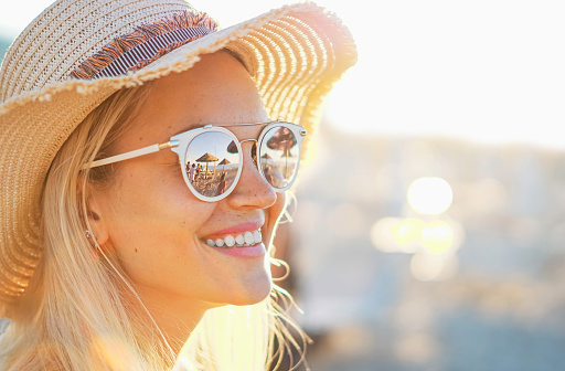 A blonde girl smiling with hat on - Beach and sunlight reflected in sunglasses - Beautiful young women in holidays - Portrait photo