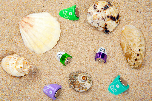 Stock photo showing close-up view of a metal bucket, pile of seashells and starfish besides a sandcastle decorated with a red triangular flag, on a sunny, golden sandy beach with sea at low tide in the background. Summer holiday, tourism and activities concept.