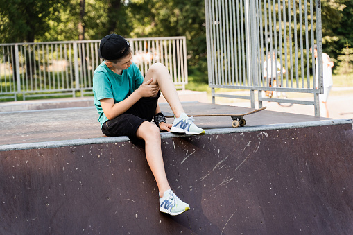 Active child boy after fall from skate board injured, sitting and looking at bruise on sport ramp on skate park playground