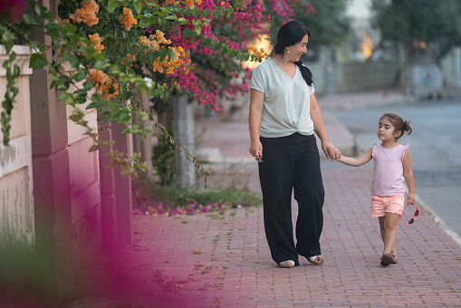 Photo of 3,5 years old girl holding mother's hand and walking on sidewalk in outdoor. Pink bougainvillea flowers are seen around. Shot under daylight with a full frame mirrorless camera.