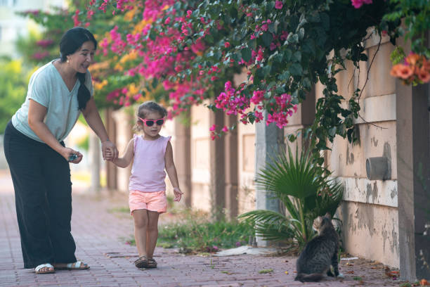 Photo of 3,5 years old girl holding mother's hand and walking on sidewalk in outdoor. Pink bougainvillea flowers are seen around. Shot under daylight with a full frame mirrorless camera.