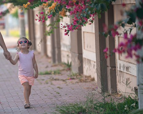 Photo of 3,5 years old girl holding mother's hand and walking in outdoor. Pink bougainvillea flowers are seen around. Shot under daylight with a full frame mirrorless camera.