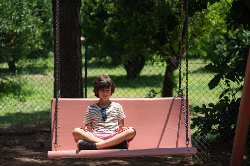 Photo of 9 years old sittin on large swing in lotus position and meditating in public park. Trees are seen on the background. Shot under daylight with a full frame mirrorless camera.