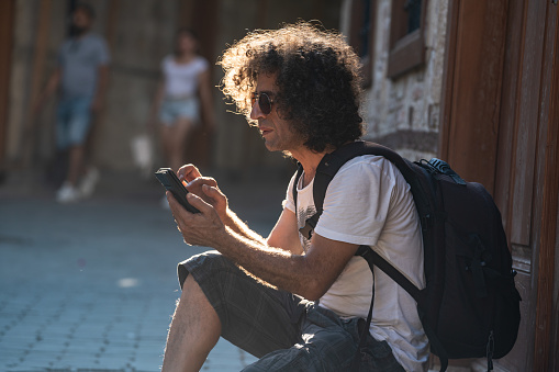 Portrait of adult man with long curly hair sitting on sidewalk and using smart phone in Old Town of Antalya, that is Kaleici, Turkey. He is wearing a white t-shirt and carrying a bag. Shot in outdoor with a full frame mirrorless camera.
