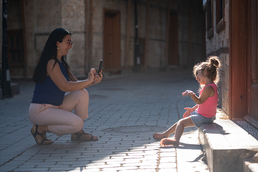 Full length photo of mother photographing her 3,5 years old daughter who is sitting on sidewalk in Old Town, Antalya, Turkey . Mother is sitting on knees and using a smart phone for photographing. Shot under daylight with a full frame mirrorless camera.