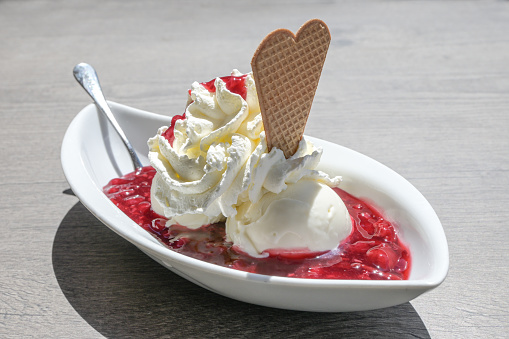 Ice cream with whipped cream, wafer cookie and red fruit jelly, in Germany called rote gruetze, served in a white bowl on a gray table on a sunny summer day, copy space, selected focus