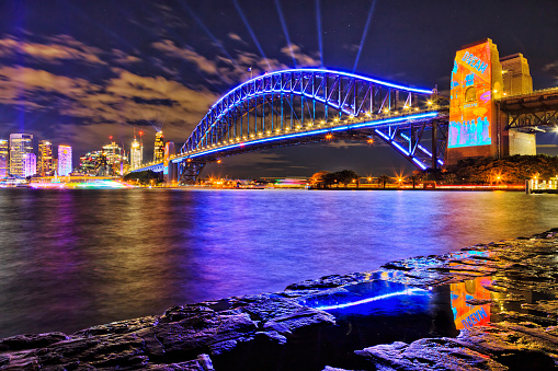 Bright blue light beams over Sydney harbour bridge with reflection in water of Harbour during Vivid Sydney festival, Australia.