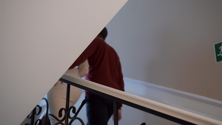 Romantic happy couple go upstairs indoors. Woman go holding palm hand on railing and carry bouquet of flowers. Side view. Rear view. Close-up.