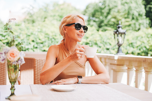 Adult elegant blonde woman in sunglasses beige clothes with cup of coffee in summer city street cafe
