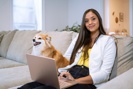 Young latin woman on the couch using computer with her dog besides