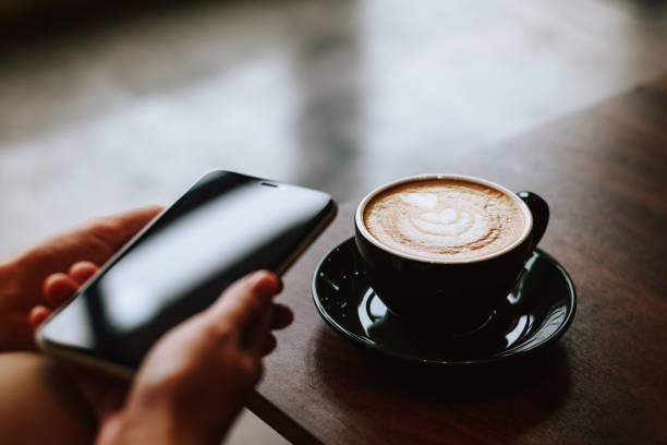 Shallow depth of field shooting of neat female hand holding plate with cup on it with delicious coffee on wooden table surrounded by another cup, glass teapot and smartphone in defocused foreground