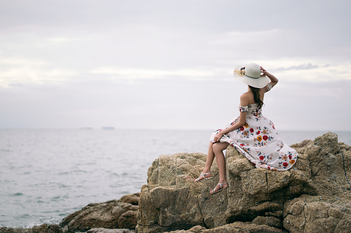Summer holiday concepy. Rear view of young adult asian woman sitting on the coast beside the sea beach. Her wear flower dress with hat.