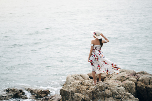 Summer tourism at tropical island concept. Rear view of young adult asian woman standing on the coast beside the sea beach. Her wear flower dress with hat luxury style pretty girl.