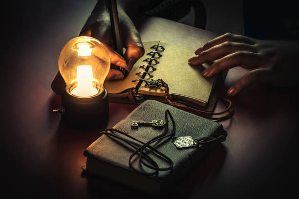Young man writing on a blank page of a vintage leather book under the low light condition with a yellow, light bulb. stock photo
