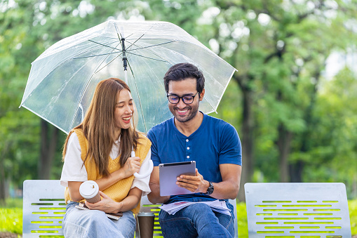 Young Asian and Indian couple enjoy having relaxing time in the rain together in the public park while sitting together on the bench during the weekend