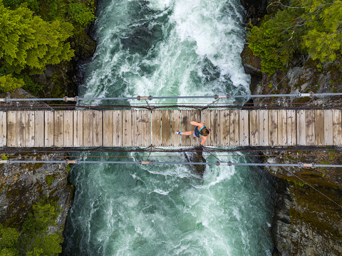 Drone view of a lush green coastal forest with a woman running over a suspension bridge. Fit female keeping active and fit. Cheakamus River in Whistler, Canada.