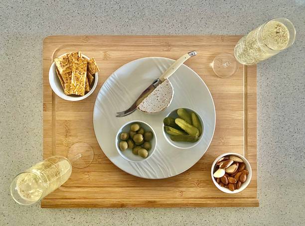 Small Appetiser Plate with Champagne Horizontal flat lay of small simple appetiser platter of cheese olives gerkins nuts and crackers on wood cutting board with champagne flutes on kitchen table appetiser stock pictures, royalty-free photos & images