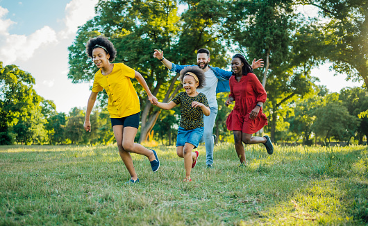 Playful multiracial family is running outside on a grassy field. They have fun while chasing each other