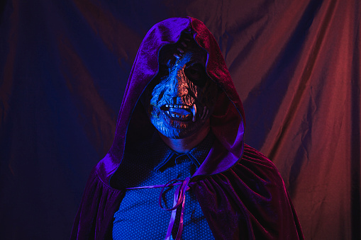 Portrait of a zombie dressed in a shirt and hooded cape facing the camera. The scene is dark, illuminated by blue and orange lights.