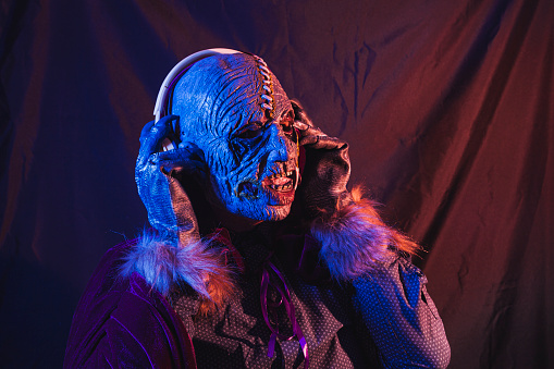 Portrait of a zombie dressed in a shirt and cape listening to music with wireless headphones. The scene is dark, illuminated by blue and orange lights.