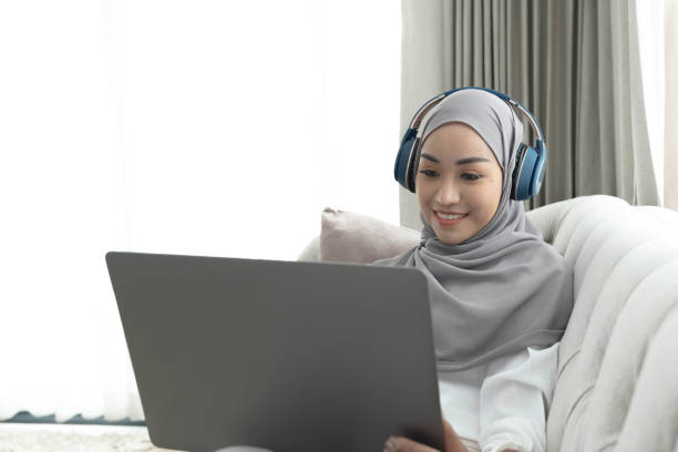 istock Charming young Asian Muslim woman wearing hijab sits on a comfy sofa, using a laptop computer and listening to music on her headphones. 1409286452