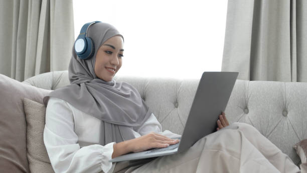 istock Charming young Asian Muslim woman wearing hijab sits on a comfy sofa, using a laptop computer and listening to music on her headphones. 1409286463