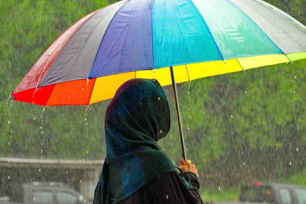 Malaysia. Sandakan. A girl in national Muslim clothes under a torrential tropical rain walks with an umbrella in rainbow colors along the coast of the island of Borneo.