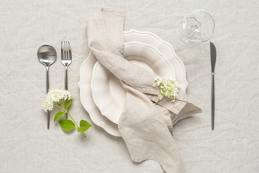 Wedding table setting. Beige plates, a beige linen napkin and white hydrangea flowers on a beige linen tablecloth. Top view, flat lay.