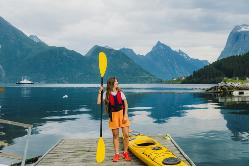 Potrait of smiling female with long hair staying on the wooden pier near yellow kayak and preparing for paddling on the fjord witth view of the beautiful mountains