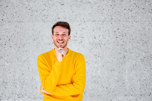Portrait of  young cheerful man in a yellow sweater standing outdoors by the concrete wall and touching his chin while looking at camera.