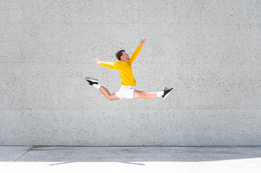 Male dancer in a yellow sweater and white shorts doing a high jump in front of a big concrete or stone wall outdoors.