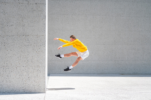 Side view of a young man wearing yellow sweater, white shorts and black sneakers, jumping by the big concrete wall like he is puhed away from it.
