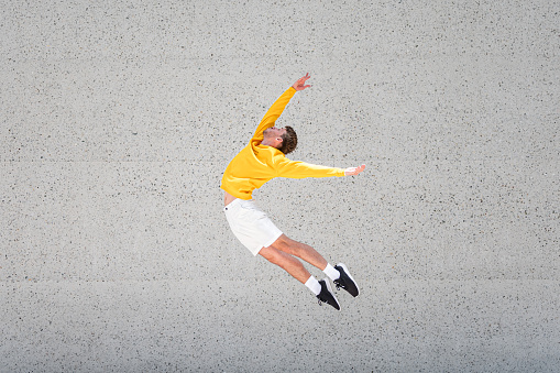 Male dancer in a yellow sweater and white shorts doing a high jump in front of a big concrete or stone wall outdoors.