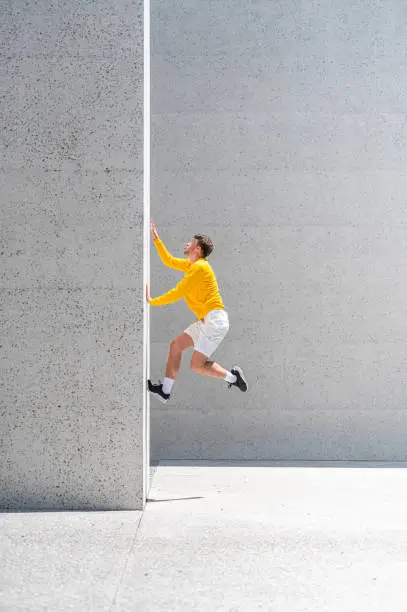 Young adult athlete wearing white shorts, yellow sweater and black sneakers jumping on the big grey concrete wall outdoors on a sunny summer day.