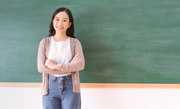 教室の真っ白な黒板の前でカメラを見つめる腕を組んで立っている美しいアジア人教師の肖像画。学校に戻るコンセプト、コピースペース付き - professor teacher female blackboard ストックフォトと画像
