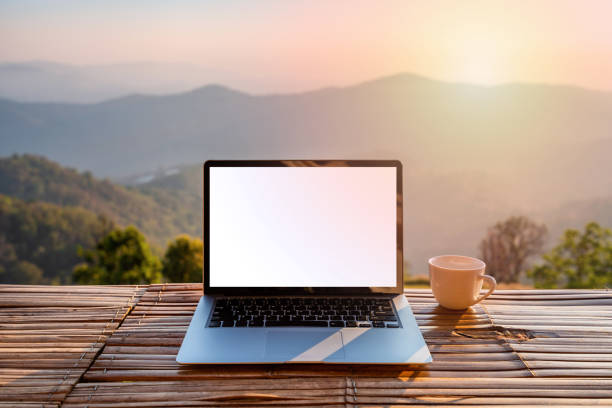 Empty screen laptop with cup of coffee and camera with mountain view at sunrise in the morning stock photo