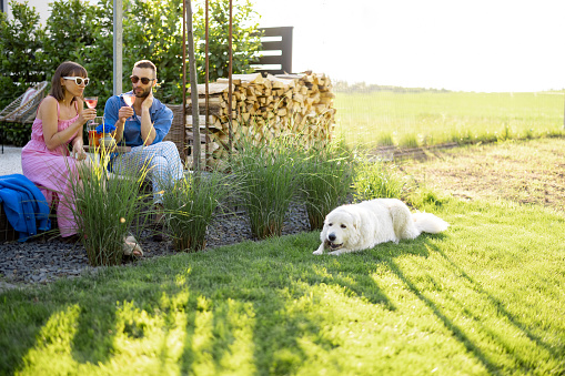 Young stylish couple have a drink while sitting together with dog on a lawn at their beautiful backyard of country house. Young man and woman spend summer time