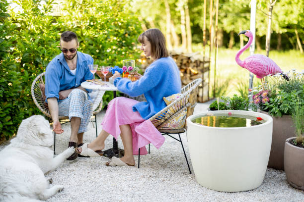 Young stylish couple have conversation while sitting by the round coffee table with dog at their beautiful garden. Young man and woman have romantic evening outdoors