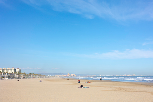 Summer vibes on the sunny autumn beach of Malvarrosa in Valencia, Spain. Vast expanses of smooth fine sand on the sea coast attract vacationers to solitary walks along the bubbling foamy waves.