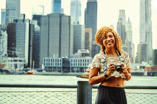 Portrait smiling young adult photographer millennial tourist woman with blond afro hair holding retro camera talking photos outdoors with Manhattan New York City skyline skyscraper behind Hudson river