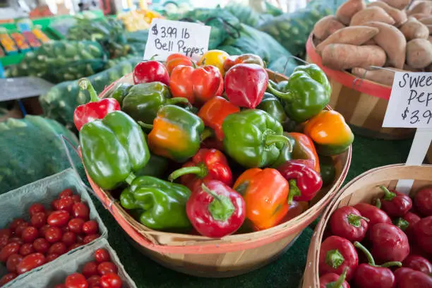 A basket of colorful bell peppers, with grape tomatoes and sweet potatoes, on display at a farmer's market
