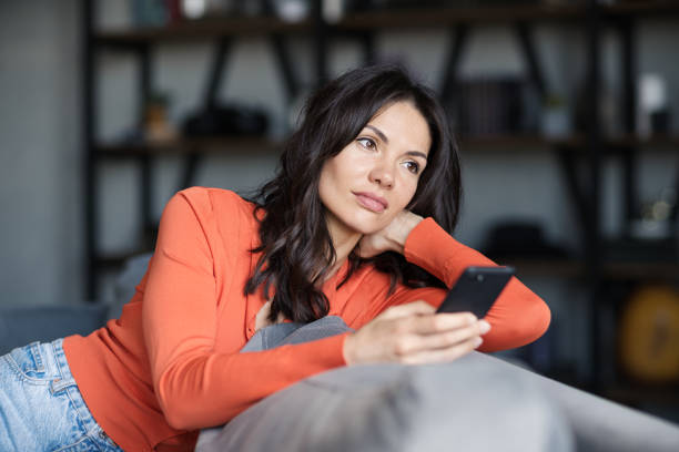 istock Attractive brunette woman sitting on sofa at home and thinking while text messaging on cell phone. Beautiful female contemplating over sms on mobile phone 1409312780