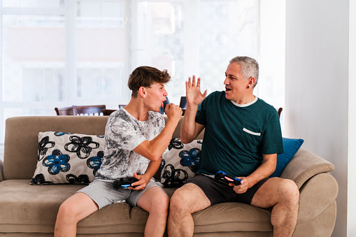 Leisure With Dad. Cheerful Father And Son Competing With Each Other In Video Games, Using Joysticks, Having Fun At Home, Free Space