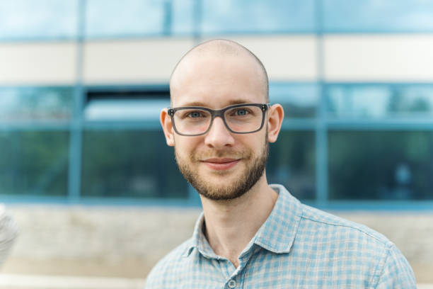 istock Intelligent young man with shaved head, beard and eyeglasses looking at camera standing outside modern business building and smiling slightly. Career, human resources and professional success concept 1409315977