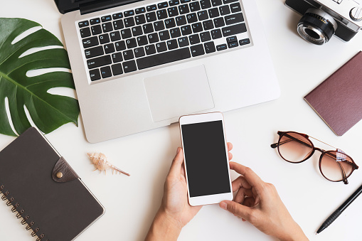 Flat lay of woman hands using mobile phone with travel item and laptop on desk. Summer, holiday, work and planning travel concepts. Top view and copy space