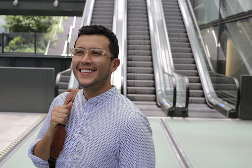 A hispanic businessman with a backpack is using a moving staircase (escalators). He looks stylish and cheerful.