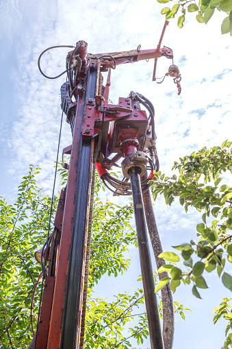 Photo of a drilling rig with a pipe for drinking water drilling against the background of trees and sky with clouds of warm sunny day.
