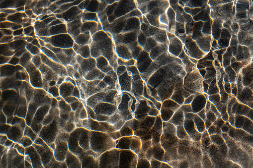 The small waves on the surface of the mountain stream bend the sun's rays. This creates bright light lines. In these patterns, the light is additionally refracted and rainbows are created over the sandy streambed.