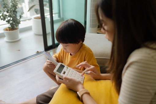 Asian pre-school boy interrupt his mother while she planning home budget in living room on weekends.