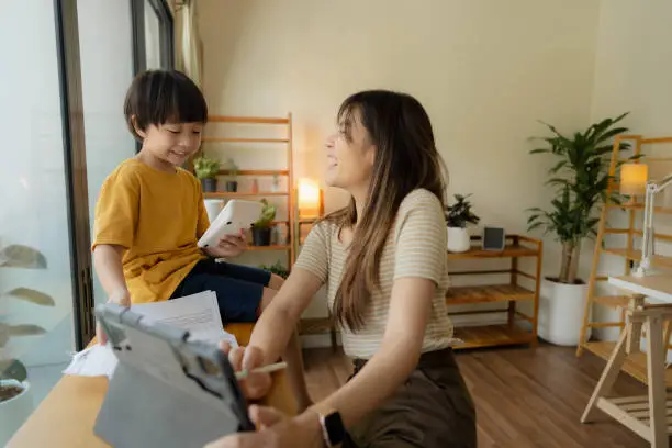 Young Asian mother sitting with pre school son in living room, holding bills and calculating home finances for savings before school starts.